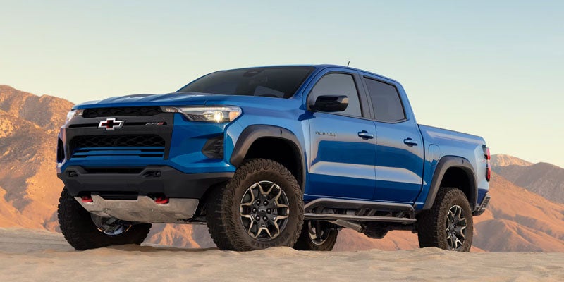 A blue pickup truck is parked on sandy terrain with mountains in the background