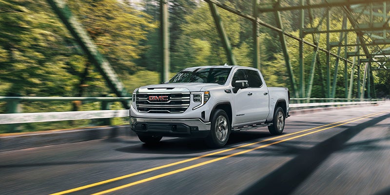 A white GMC pickup truck is driving across a metal bridge surrounded by green trees