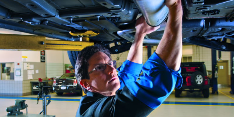 A person in a workshop uses both hands to work on the underside of a raised vehicle