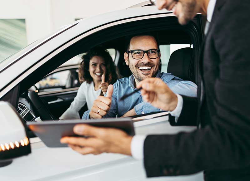 A person inside a vehicle gives a thumbs-up gesture while another person sits beside them